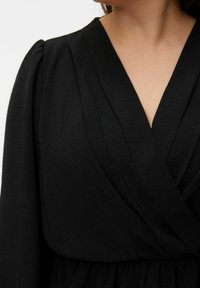 Robe portefeuille noire avec col en V, en tissu texturé. Manches longues et taille froncée. Apparence lisse et fluide.