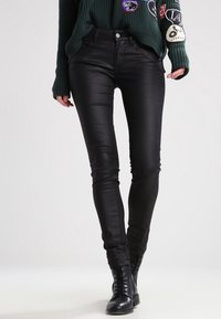 Jeans negros ajustados hechos de una tela lisa y elástica. Presenta una cintura de altura media, cinco bolsillos y un ajuste elegante. Combinados con un suéter verde texturizado.
