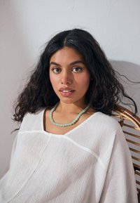 Woman with long dark curly hair sits on a wicker chair wearing a white textured blouse and a pale green beaded necklace.