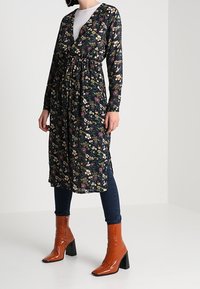 Blommig omlottklänning med långa ärmar, svart bas och färgglada blomsterupptryck. Betoningen vid midjan med knytning, ihopparad med glänsande orange klackstövlar.