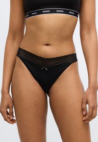 Crne tange s čipkastim rubom na struku, s valovitim rubovima i malim mašnom u sredini. Mekani tekstura tkanine.