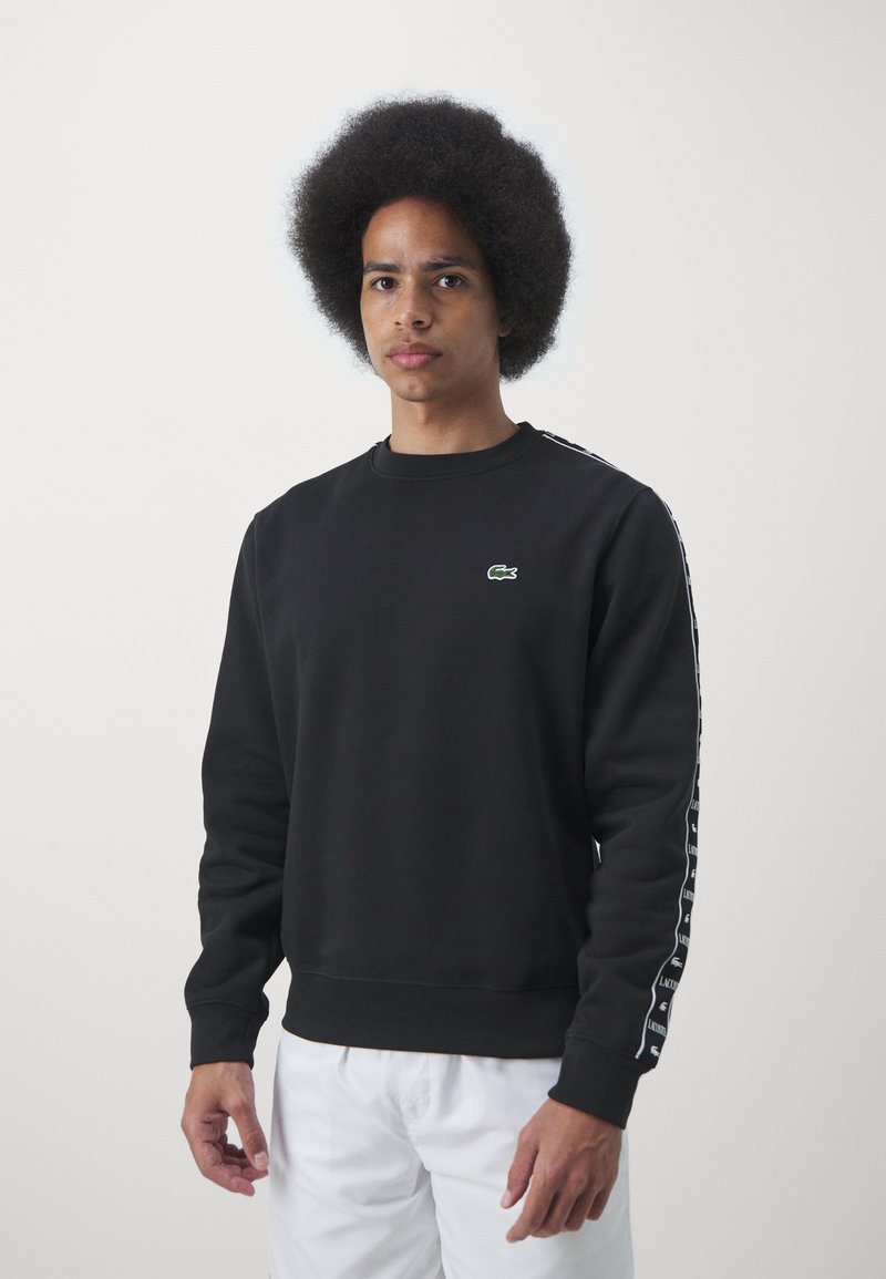 Schwarzer Sweatshirt mit Rundhalsausschnitt, langen Ärmeln und weißen Seitenstreifen mit Branding. Grünes Logo auf der linken Brust. Hergestellt aus strukturiertem Stoff.