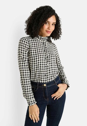 Femme aux cheveux bouclés portant une blouse à carreaux noir et blanc et un jean bleu foncé, posant avec une main dans sa poche.