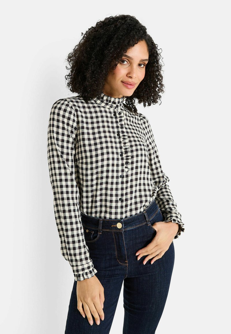 Mujer con cabello rizado que lleva una blusa a cuadros en blanco y negro y jeans de color azul oscuro, posando con una mano en el bolsillo.