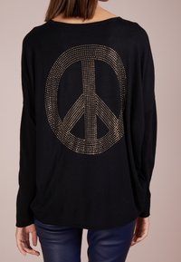 Svart tröja med en stor guldnubbad fredssymbol på ryggen. Designen har en lös passform med långa ärmar och en rundad halsringning.