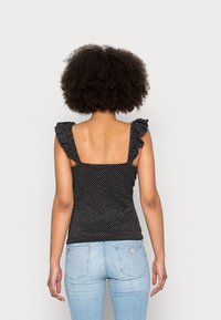 Schwarzes ärmelloses Oberteil mit weißen Punkten, Rüschen an den Trägern, figurnahe Passform und quadratischem Rückenteil, getragen mit hellblauen, eng anliegenden Jeans.