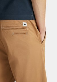 Pantaloni khaki con tasche posteriori e etichetta bianca con un logo nero. Materiale in cotone, design diritto, texture liscia e vita aderente.