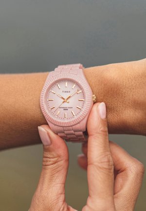 Timex horloge met een roze siliconen band en wijzerplaat, met gouden accenten op de uurmarkeringen en wijzers, een getextureerde lunette en een waterbestendig ontwerp.
