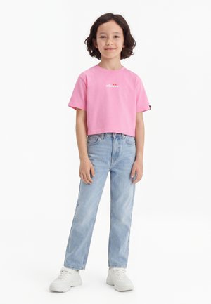 Bambino in piedi, indossa una maglietta rosa corta con logo bianco, jeans azzurri chiari e scarpe da ginnastica bianche su uno sfondo bianco semplice.