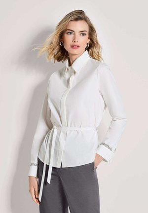 Weißes Langarmhemd mit Kragen, gebundener Taille und Verzierungen an den Manschetten. Getragen mit grauer Hose, glatte Stofftextur.