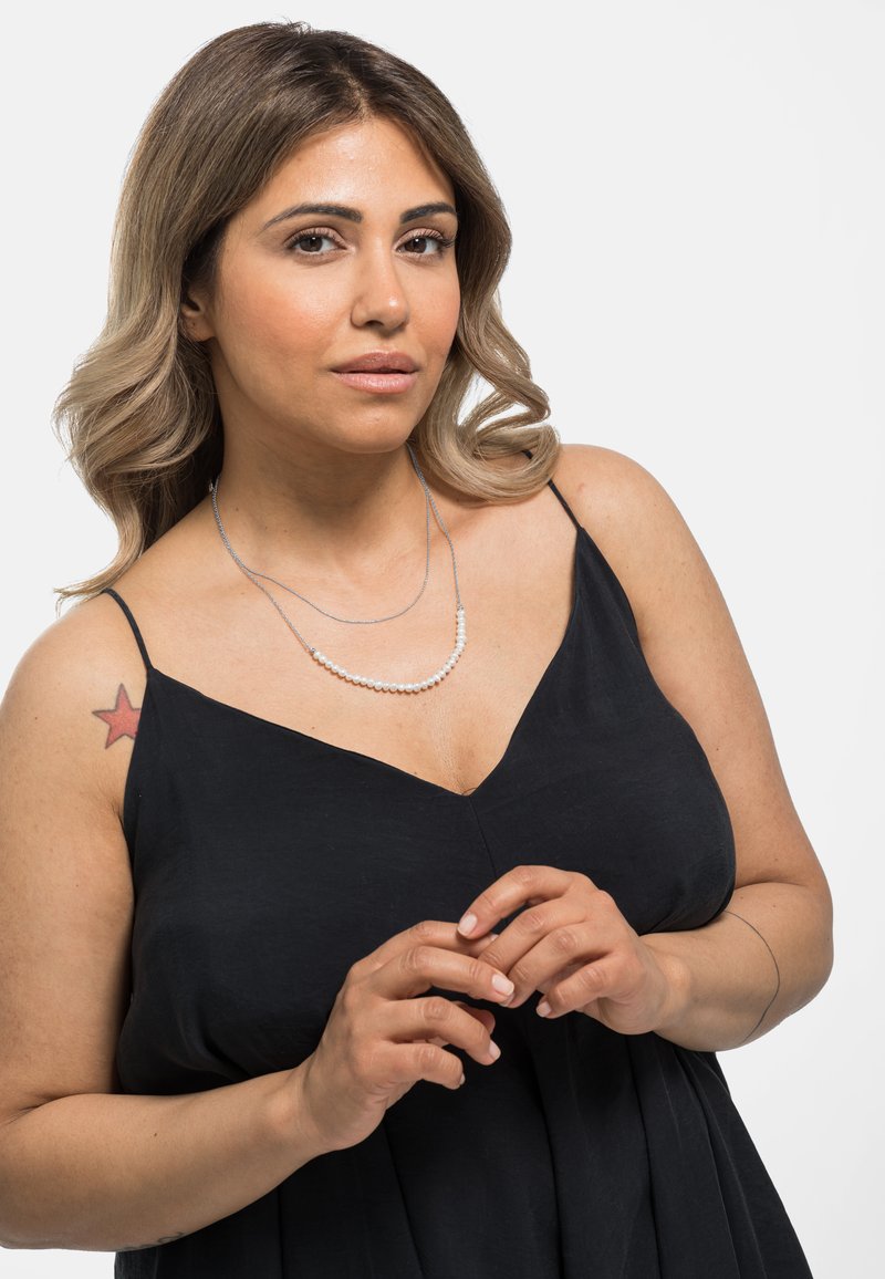 Dubbele ketting met zilveren kettingen en kleine witte kralen. Het onderwerp draagt een zwart mouwloos jurkje, waarop een ster tattoo op de schouder zichtbaar is.