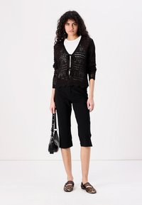 Melna adīta džempera kardigāns ar jostu virs baltās blūzes, saskaņots ar melniem svaru biksēm un leopardu raksta dzīvokļiem. Melna soma rokā.