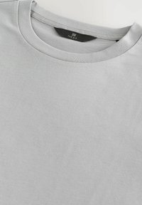 Lichtgrijze crewneck t-shirt van zachte katoenstof met een gladde textuur. Heeft een minimalistisch ontwerp en een zwart merklabel op de hals.