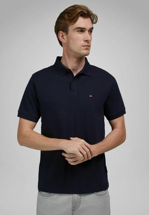 Homme aux cheveux châtains clairs portant un polo bleu marine et un jean gris clair, debout les mains jointes, regardant sur le côté.