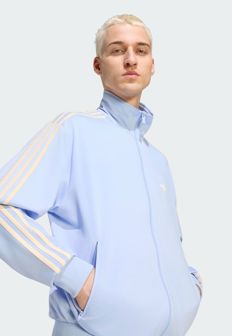 Young person with short blond hair wearing a light blue zip-up jacket with beige stripes and hands in pockets.