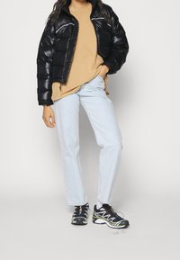 Black puffer jacket, beige crewneck shirt, light blue straight-leg jeans, and black sneakers with green accents and white details.