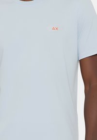T-shirt azzurro chiaro a maniche corte realizzato in tessuto morbido. Presenta un piccolo logo arancione sul lato sinistro del petto. Collo a giroclassico.