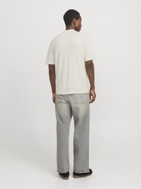 T-shirt beige clair à manches courtes, avec une coupe décontractée, associé à un jean large délavé et des chaussures noires, vu de dos.