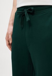 Joggers verde scuro con una texture morbida, dotati di una vita con cordoncino e una tasca laterale. Il tessuto appare traspirante e informale.