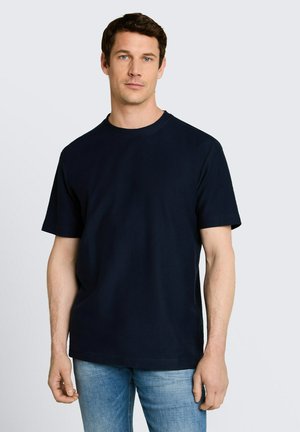 Mężczyzna o krótkich brązowych włosach, ubrany w jednolitą czarną koszulkę i niebieskie dżinsy, stojący na jasnoszarym tle.