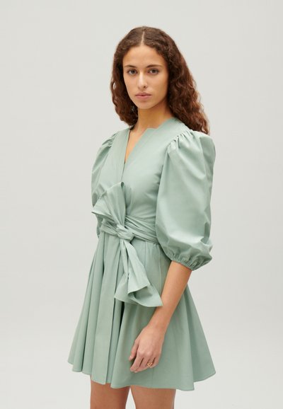 Rochie scurtă verde mentă cu mâneci pufoase, decolteu adânc în V și talie legată. Fustă pliată cu o textură fină și o țesătură moale.