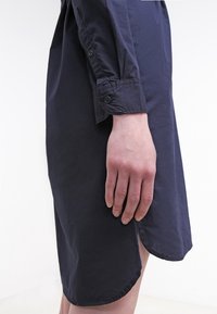 Gros plan sur la main d’une personne reposant à côté d’une robe-chemise bleu marine foncé à manches longues, avec un poignet boutonné et une fente incurvée à l’ourlet.