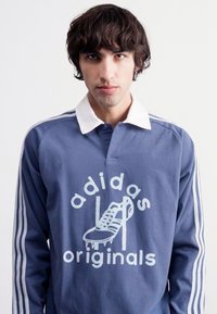 Plava duga rukava polo majica s bijelim kragnom i Adidas Originals logom koji sadrži grafiku cipela. Prugasti detalji na rukavima.
