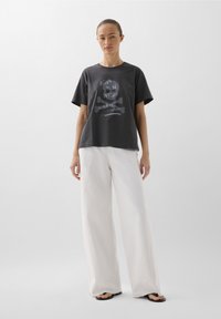 Schwarzes Baumwoll-T-Shirt mit einem Frontdrucktext "BORN TO BE WILD" und einem Totenkopfdesign, kombiniert mit weit geschnittenen weißen Hosen und schwarzen Sandalen.
