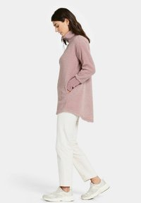 Rosa Fleece-Tunika mit hohem Kragen und Seitentaschen; lange Ärmel mit kontrastierenden Bündchen; kombiniert mit weißen Hosen und hellen Sneakern.
