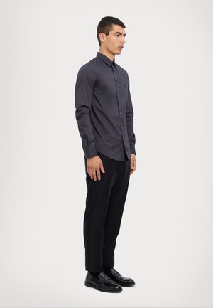 Camisa gris de manga larga con diseño abotonado, que cuenta con un collar y un único bolsillo frontal, combinada con pantalones negros y zapatos negros brillantes.
