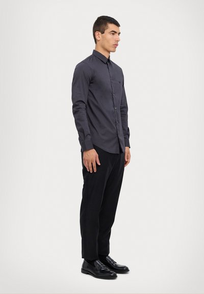 Camisa gris de manga larga con diseño abotonado, que cuenta con un collar y un único bolsillo frontal, combinada con pantalones negros y zapatos negros brillantes.