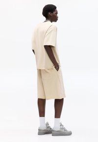 Béžové oversized tričko s krátkými rukávy, spárované s kraťasy po kolena s zadní kapsou. Šedé tenisky a bílé sportovní ponožky.