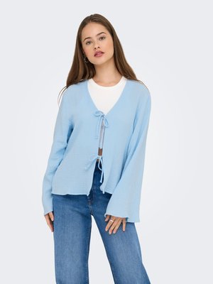 Jeune femme portant un cardigan bleu clair noué devant, avec un haut blanc et un jean bleu, debout devant un fond clair.