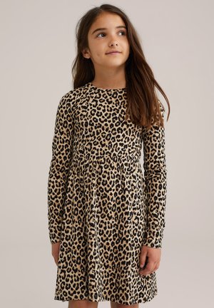 Ragazza con lunghi capelli castani che indossa un vestito a maniche lunghe beige e nero con stampa leopardata, che guarda leggermente in alto e di lato.