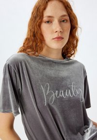 Camiseta de terciopelo gris con mangas cortas, que presenta un texto decorativo "Beauty" en un color claro que contrasta. Textura suave y corte relajado.