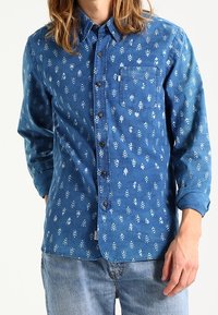 Blauwe spijkerblouse met witte bloemenpatronen, lange mouwen, een knoopsluiting aan de voorkant en een borstzak. Zachte textuur met een ontspannen pasvorm.