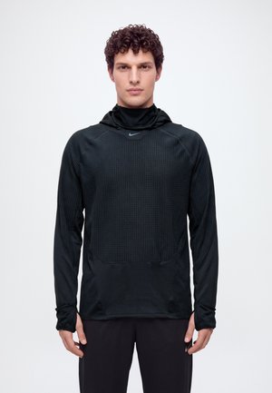 Un homme se tient face à l’objectif sur un fond blanc uni. Il porte un haut noir Nike à capuche et à manches longues, ainsi qu’un pantalon noir.