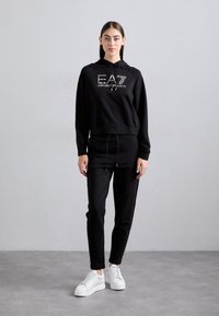Jauna moteris stovi vilkinti juodą EA7 Emporio Armani megztinį su kapuci ir atitinkančias juodas kelnes, su baltomis sportinėmis bateliais prieš pilką fono.