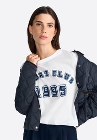 Biela tričko s modrým potlačou "SPORT CLUB 1995", v kombinácii s tmavou prešívanou bundou s textúrovaným vzorom a patentkami.