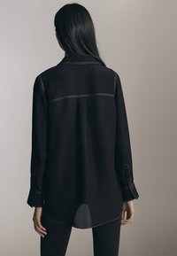 Sort skjorte med krave, lange ærmer og synlige syninger. Stofferne ser glatte ud, med en lidt løs pasform og en buet søm.
