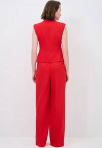 Roter taillierter ärmelloser Blazer mit Schulterpolstern, kombiniert mit weit geschnittenen Hosen. Glatte Textur, strukturierte Nähte und eine figurbetonte Silhouette.