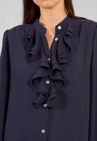Blusa blu navy con design frontale a volant, dotata di colletto rotondo e piccoli bottoni. Texture liscia con un aspetto fluido.