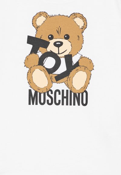 Balta grafiskā T-kreklā attēlots zīmējums ar tēju lācīšu brūnos toņos, kas tur melnu uzrakstu "TOY", ar vārdu "MOSCHINO" zem tā.