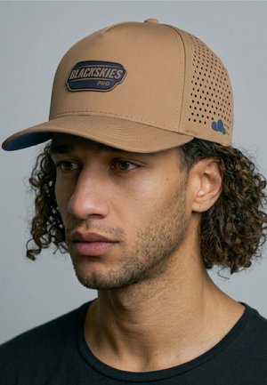 Man with curly hair wears a tan perforated cap labeled "Blackskies Pro" and a black shirt, looking slightly left against a plain background.