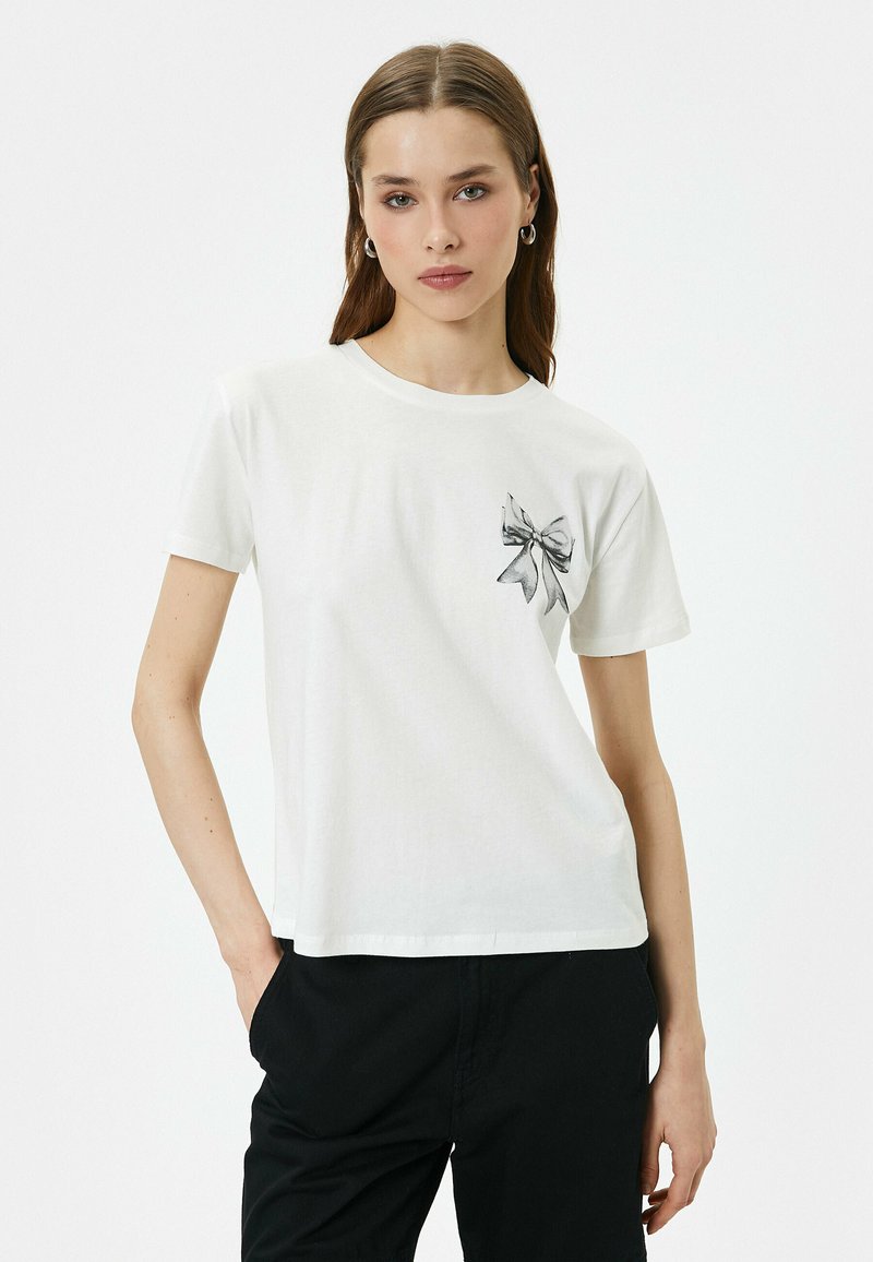 Hvid bomulds T-shirt med grafisk sort sløjfe på venstre bryst. Korte ærmer og rund halsudskæring. Glat tekstur og afslappet pasform.