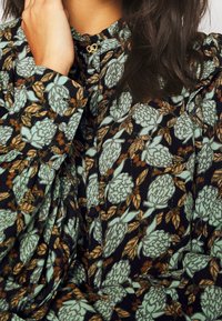 Blusa floreale con una base nera e motivi di fiori verdi e marroni, dettagli a pieghe e un gancio dorato col colletto.
