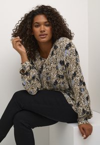 Floral blouse in beige with blue and black patterns, long sleeves, and a V-neck design. Paired with black fitted pants.