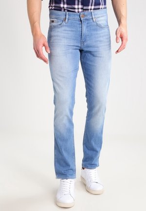 Mężczyzna ubrany w jasnoniebieskie dopasowane dżinsy, białe trampki oraz niebieską koszulę w kratę, stojący na tle jasnego, jednolitego tła.