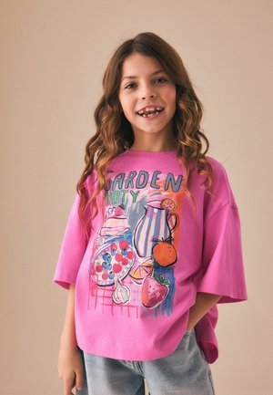 Mädchen mit langen, welligen Haaren lächelt, trägt ein übergroßes pinkes T-Shirt mit buntem "Garden Party"-Motiv und blaue Jeans, die Hände in den Taschen.