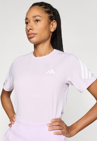 Jasnofioletowa sportowa koszulka z krótkim rękawem, okrągłym dekoltem i białymi paskami na rękawach. Na piersi znajduje się małe logo marki.